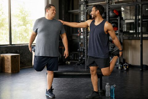 Dos hombres adultos con cuerpos distintos se ríen mientras hacen un estiramiento sencillo en un gimnasio