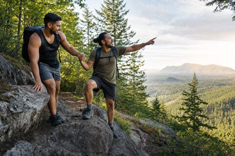 Dos hombres adultos se ayudan en un sendero; uno ofrece apoyo y el otro señala la ruta entre las montañas